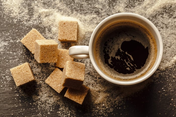 Dark coffee in white cup beside scattered brown sugar cubes and granules