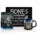 Two twelve-ounce bags of High Voltage coffee are in front of a black box that says Bones Coffee Company. Beside the box is the High Voltage handthrown mug that is black colored with a blue glaze on top of it. The mug’s medallion has the art of the High Voltage coffee on it.
