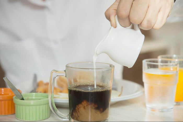 person pouring milk into cup of black coffee on breakfast table