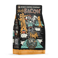 The front of a 12 ounce bag of Bones Coffee Company Maple Bacon coffee. Its flavor is bacon, and it has a skeleton dressed like a lumberjack sipping coffee in a forest with pigs on the art.
