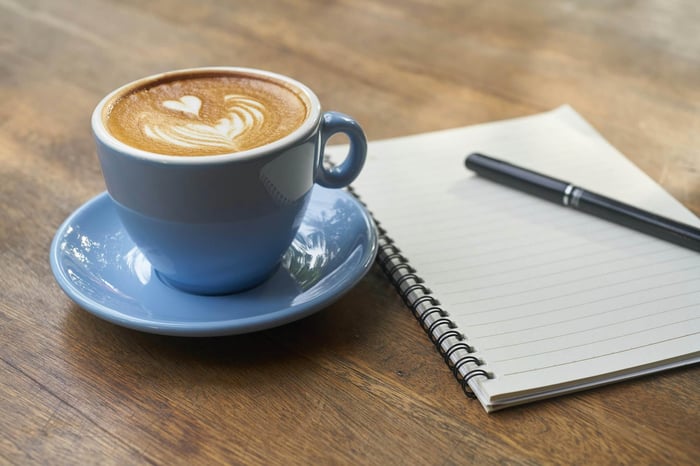 Blue coffee cup with heart latte art beside a notebook and pen