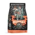 The front of a 12 ounce bag of Bones Coffee Company Rest In Peace coffee. There is a skeleton wearing a nightcap sitting on a rocking chair, reading a book and drinking coffee on the art.